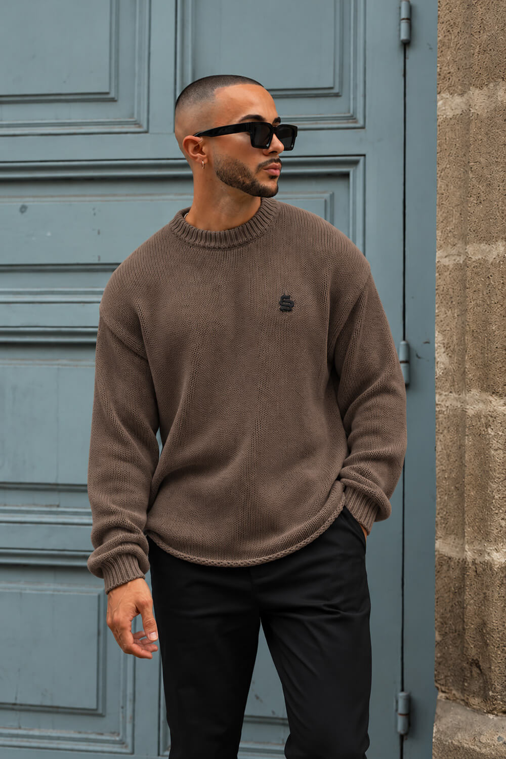A man wearing a brown knit crewneck sweater with a small black embroidered logo on the left chest, paired with black pants and sunglasses, standing in front of a blue paneled door.