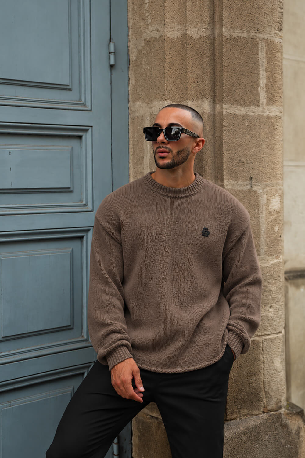 A man wearing a brown knit crewneck sweater, black pants, and black sunglasses leans against a stone wall next to a blue door.