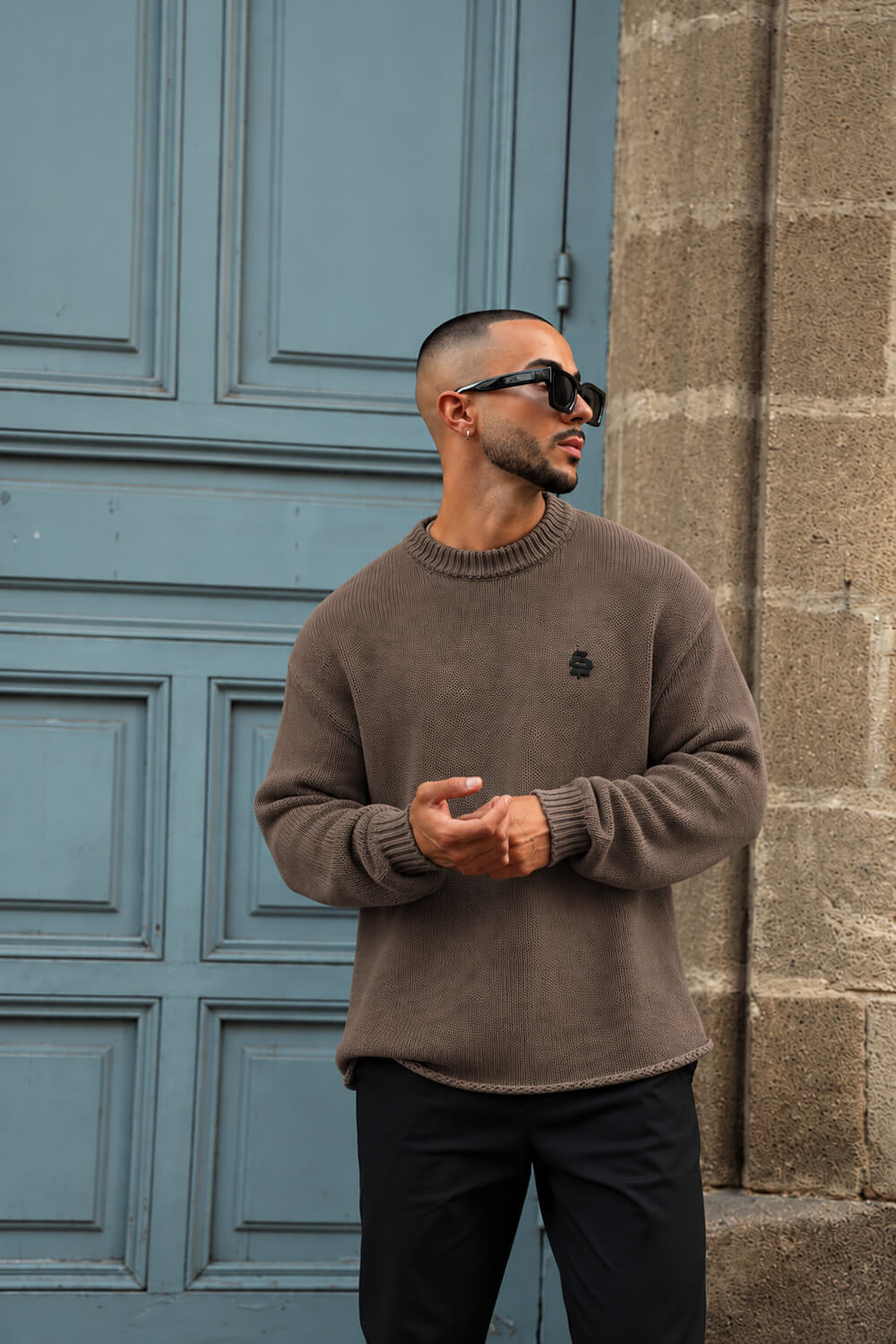 A man wearing a brown crewneck knit sweater and black sunglasses, standing in front of a blue paneled door and a stone wall.