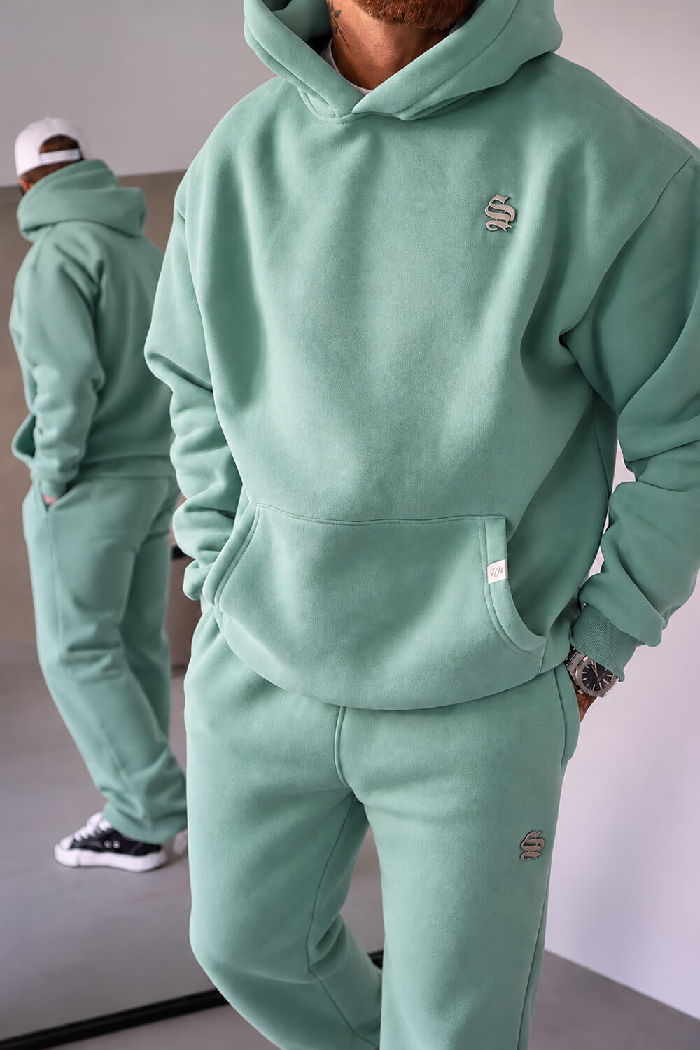 A person wearing a matching sage green hoodie and sweatpants set, with a mirror in the background reflecting the back of the outfit. The hoodie features a small embroidered logo on the left chest.