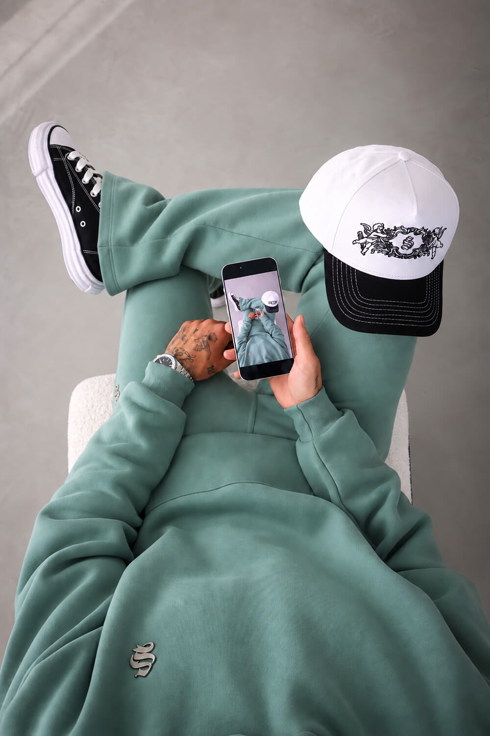 A person wearing a sage green sweatshirt and matching sweatpants, black and white sneakers, and a white and black baseball cap, looking down at a phone on their lap against a gray concrete background.