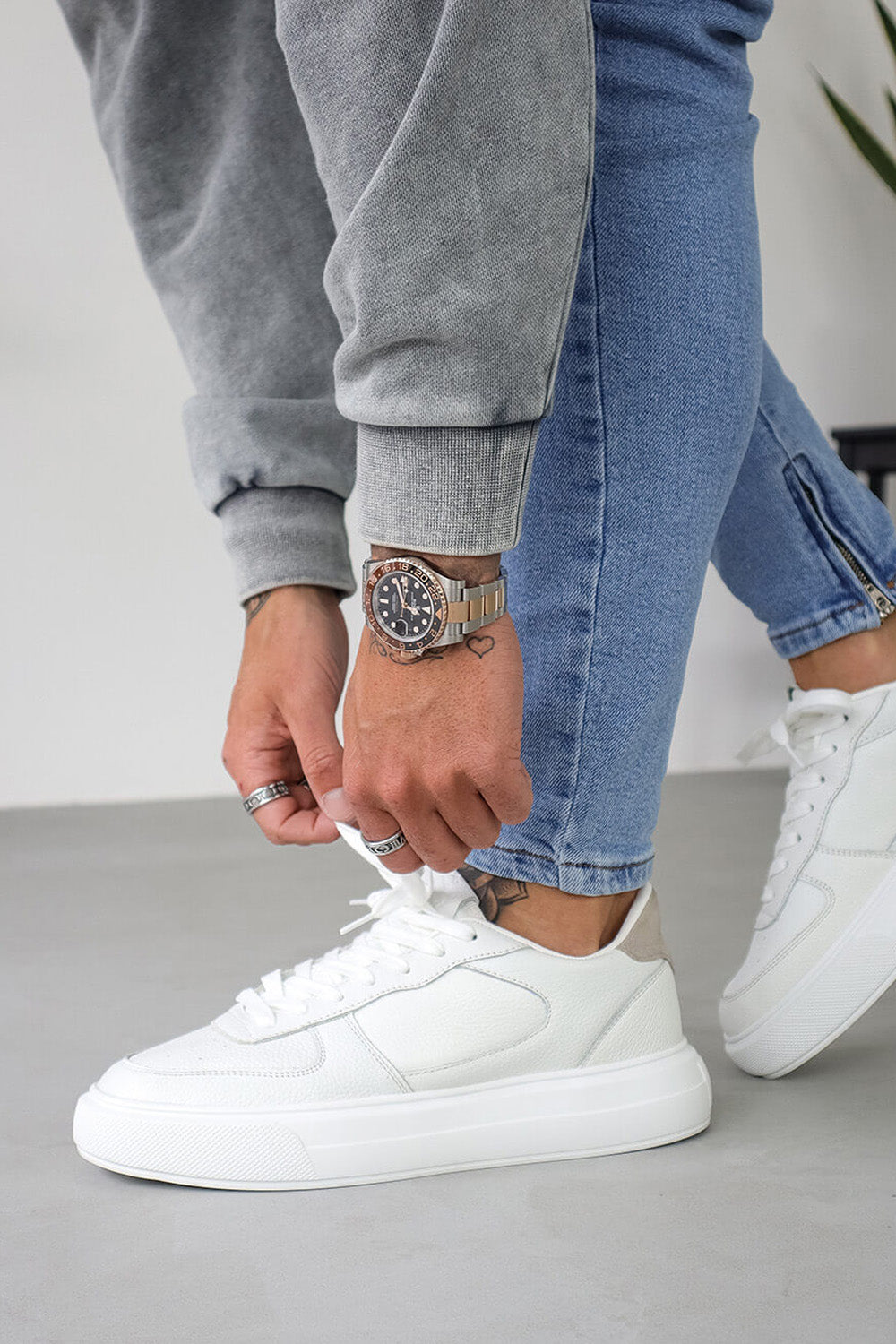 A person wearing grey sweatshirt and blue jeans, with white triple white court trainers