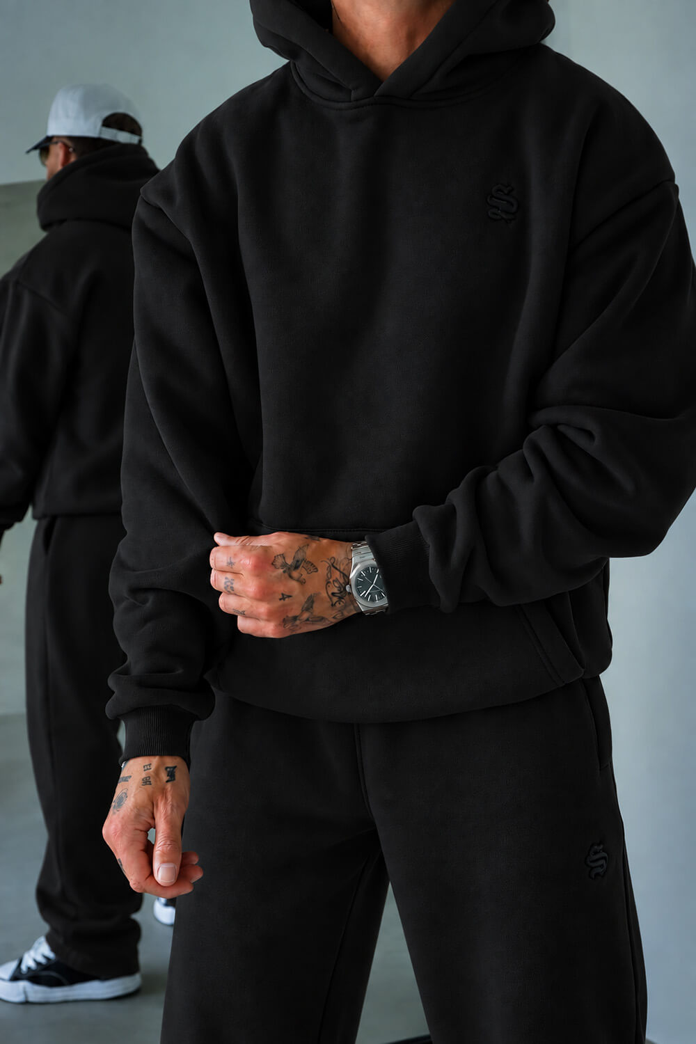 A person wearing an oversized black hoodie and matching black sweatpants, shown from the neck down in a studio setting. The hoodie has a subtle tonal embroidered logo on the left chest, and the matching sweatpants have a small tonal logo near the lower leg.