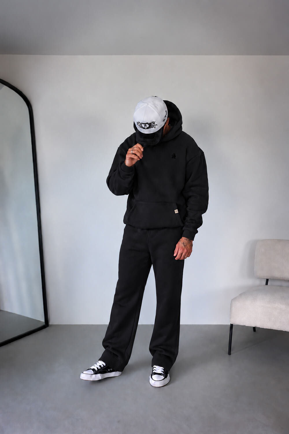 A person modeling a black hooded sweatshirt and matching black sweatpants in a minimalist room with a light gray wall, concrete floor, a large black-framed mirror on the left, and a light beige chair on the right.