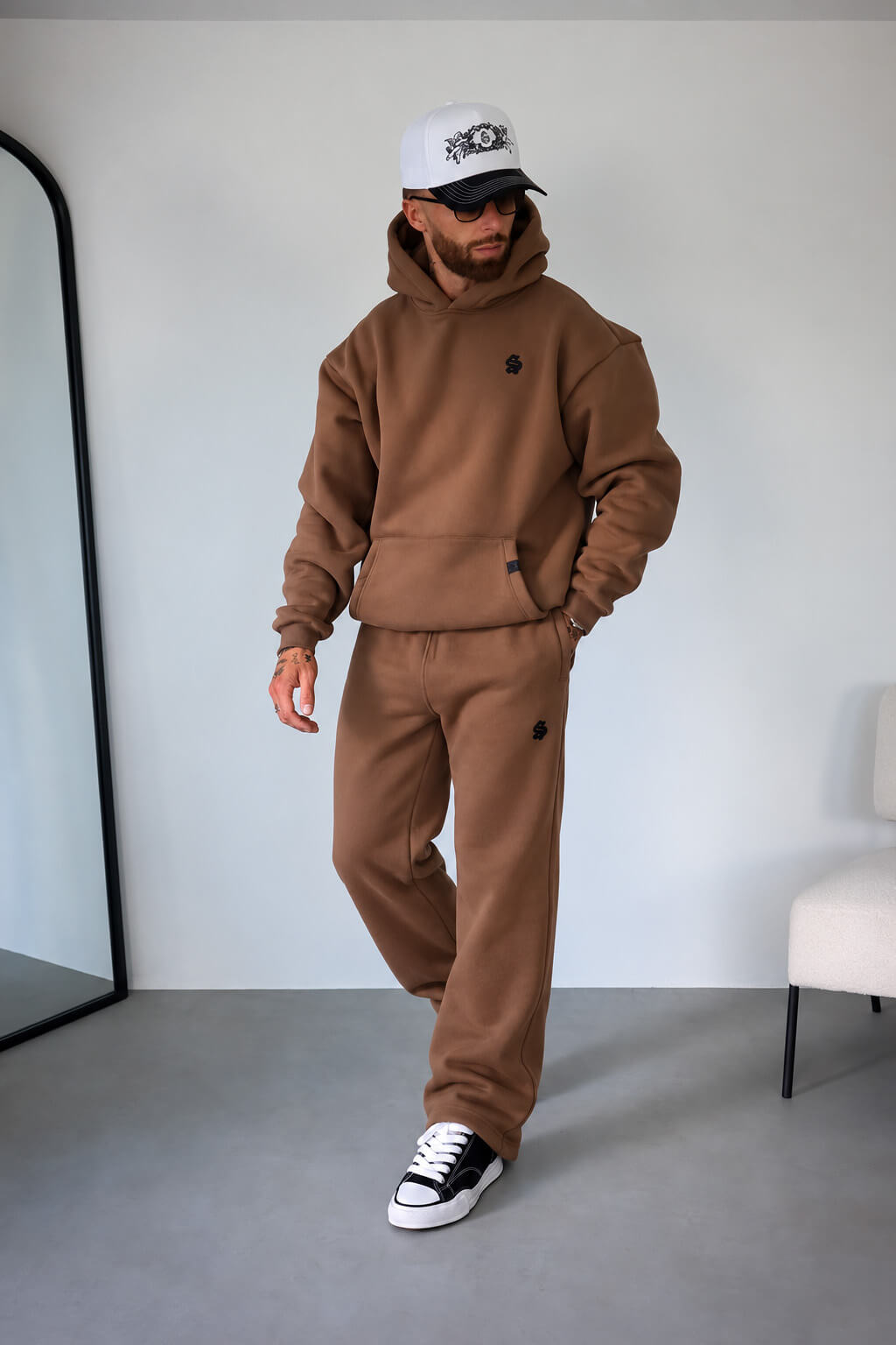 A man modeling a matching brown fleece hoodie and sweatpants set, styled with a white baseball cap, black sunglasses, and black-and-white sneakers in a minimalist room.