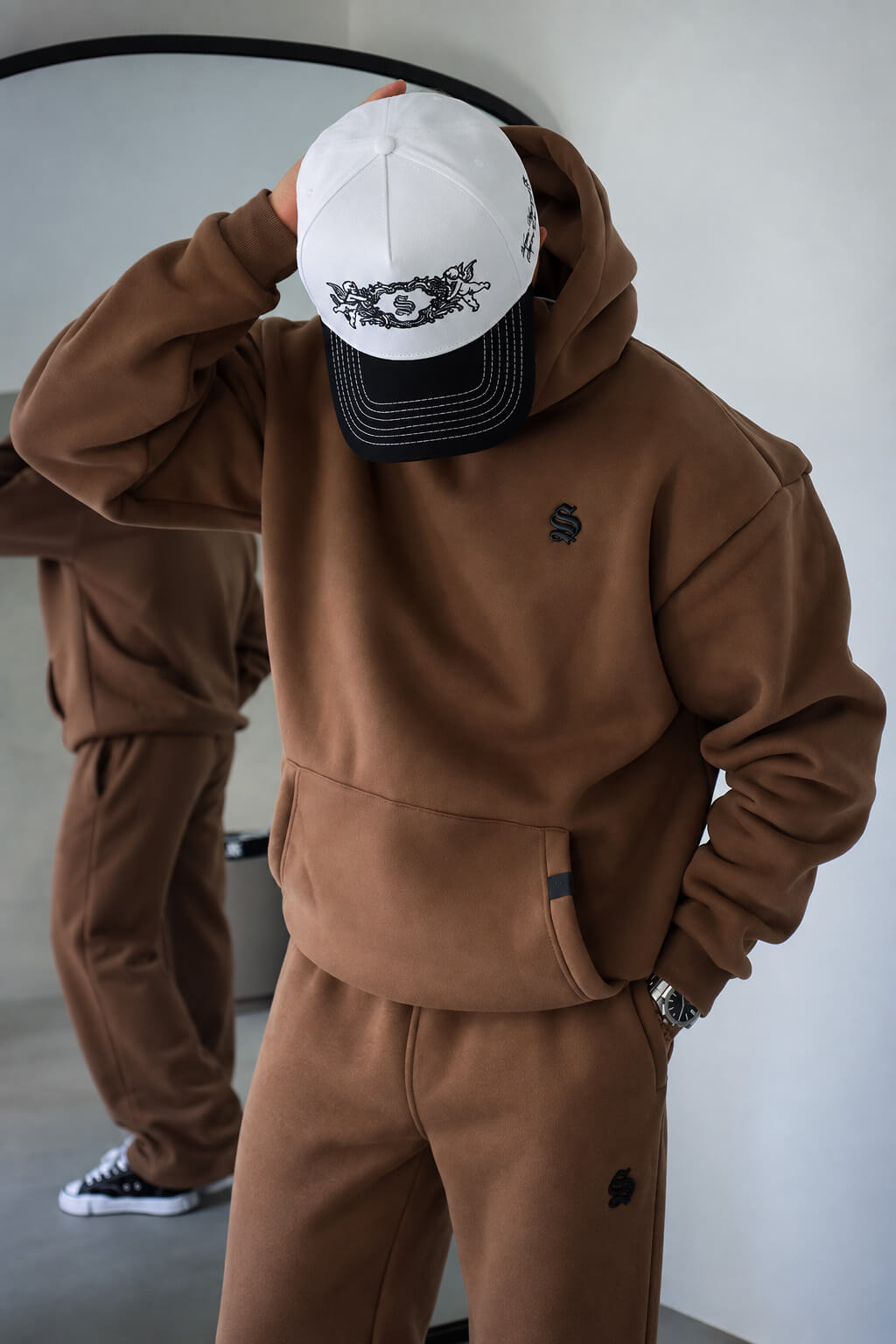 A person wearing a brown hooded sweatshirt and matching brown sweatpants, with a white and black baseball cap covering the face. The setting appears to be an indoor studio or room with a mirror reflecting the lower body.