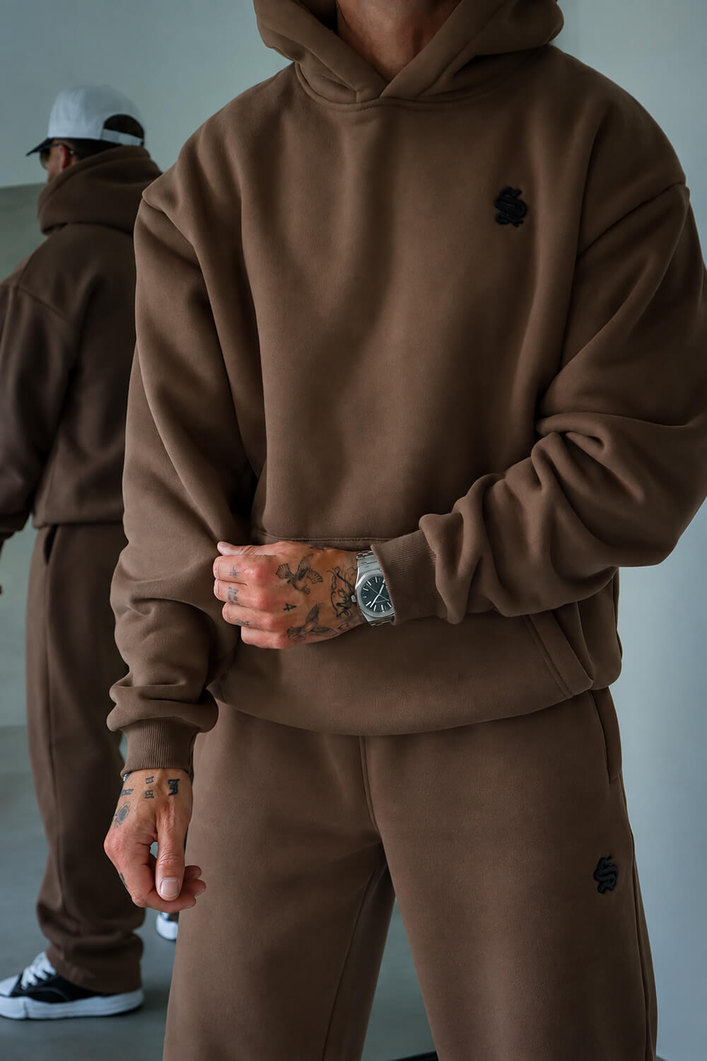 A person wearing a matching brown hoodie and sweatpants set, with a small black embroidered logo on the chest and left thigh. The photo is cropped from the neck down, showing tattooed hands and a silver watch, with another person blurred in the background wearing the same outfit.