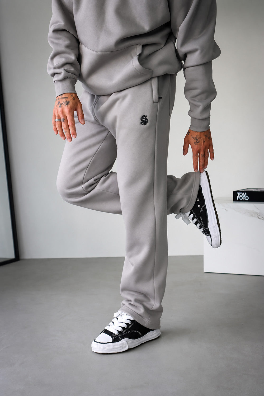 A person wearing light gray sweatpants and a matching gray hoodie, paired with black and white sneakers, posing in a minimalist gray room.