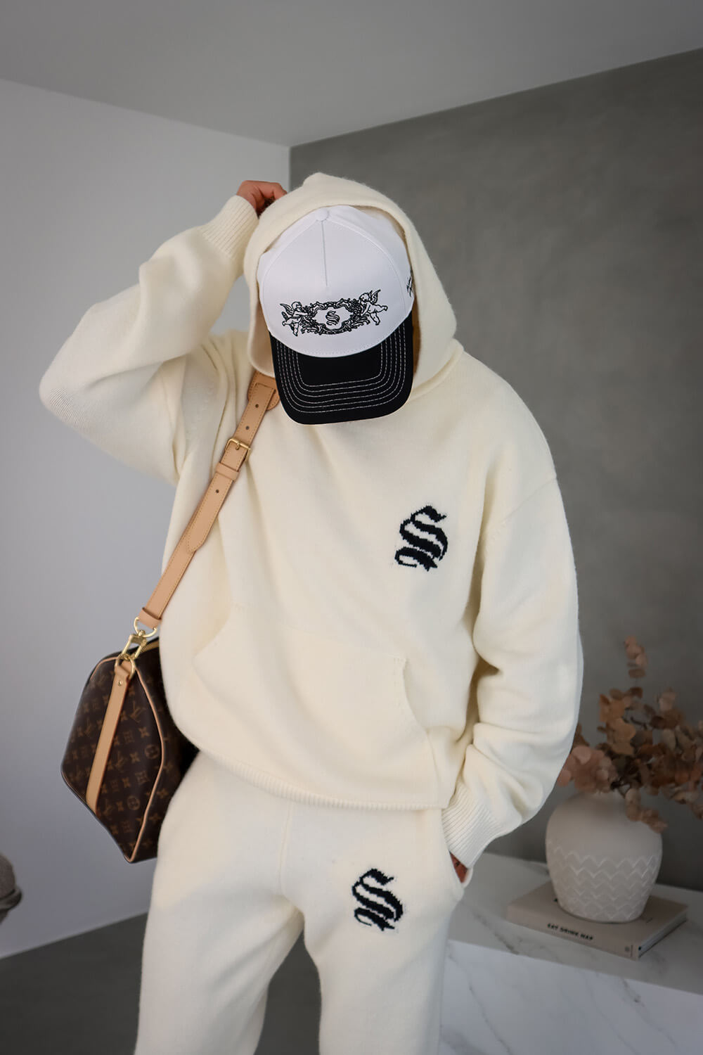 Person wearing a white hoodie and pants with a black logo, holding a white cap with black design, and carrying a brown handbag.