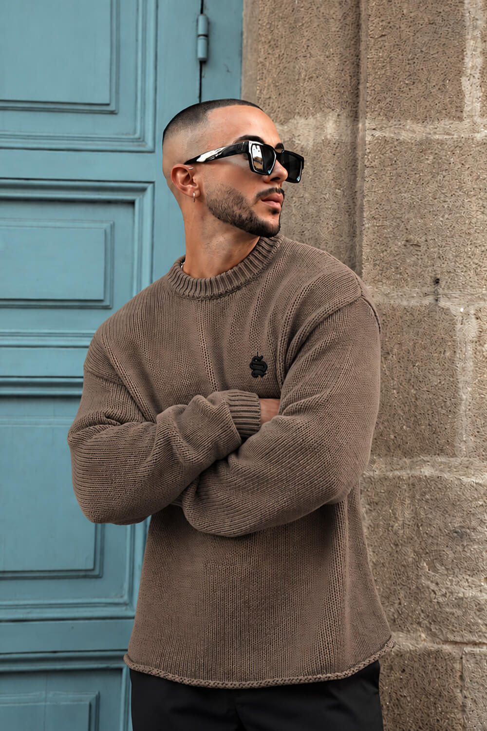 A man wearing a brown ribbed knit crewneck sweater and black sunglasses, standing with arms crossed in front of a blue door and a stone wall.