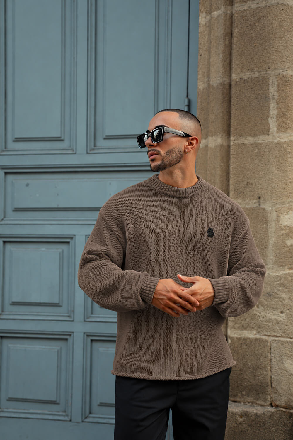 A man wearing a taupe brown knit sweater and black sunglasses stands in front of a blue paneled door and a stone wall.