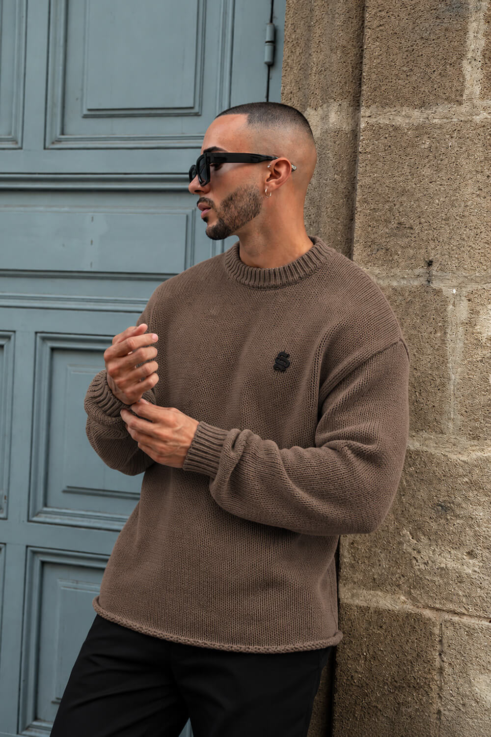 A man wearing a brown knit crewneck sweater and black sunglasses leans against a stone wall next to a blue paneled door.