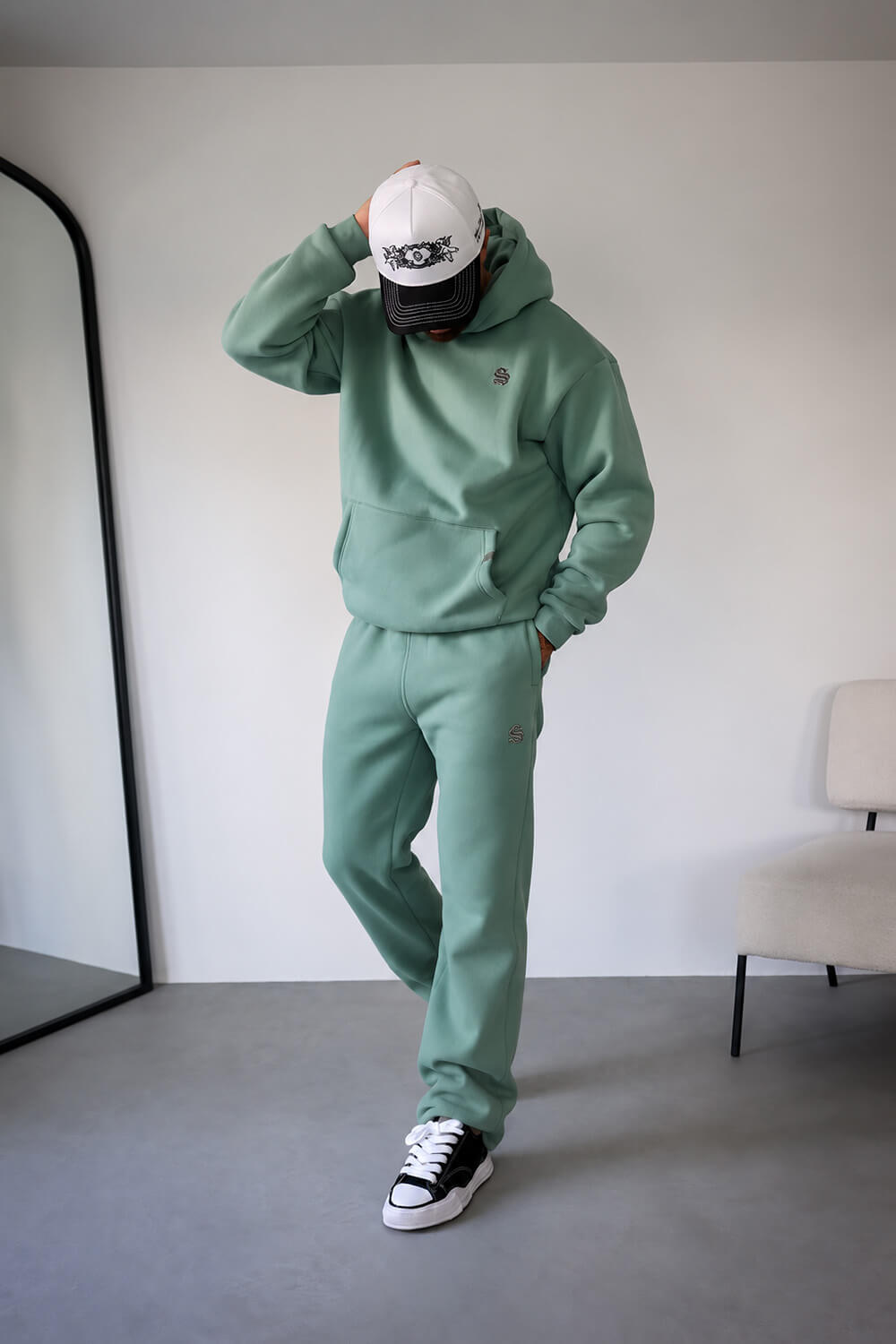 A person modeling a matching sage green hoodie and sweatpants set, paired with a white and black baseball cap and black-and-white sneakers, standing in a minimalist room with a gray floor and white wall.