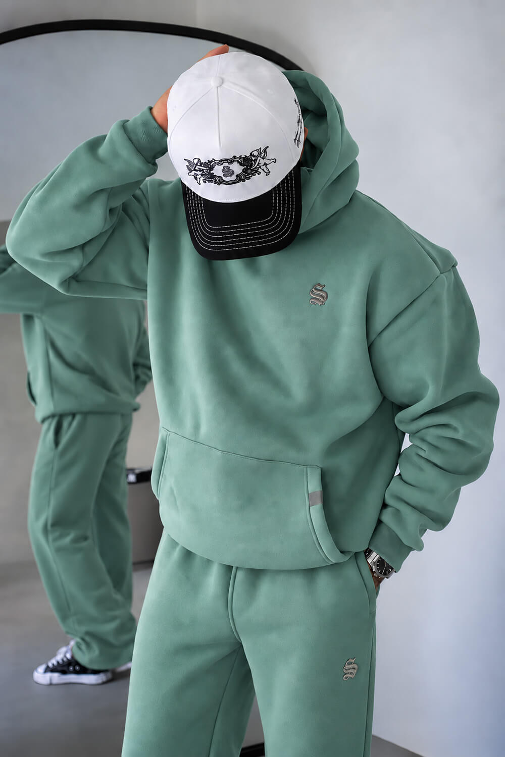A person wearing a sage green hooded sweatshirt and matching sweatpants, with a white and black baseball cap, standing indoors. A mirror in the background shows the person's full body.