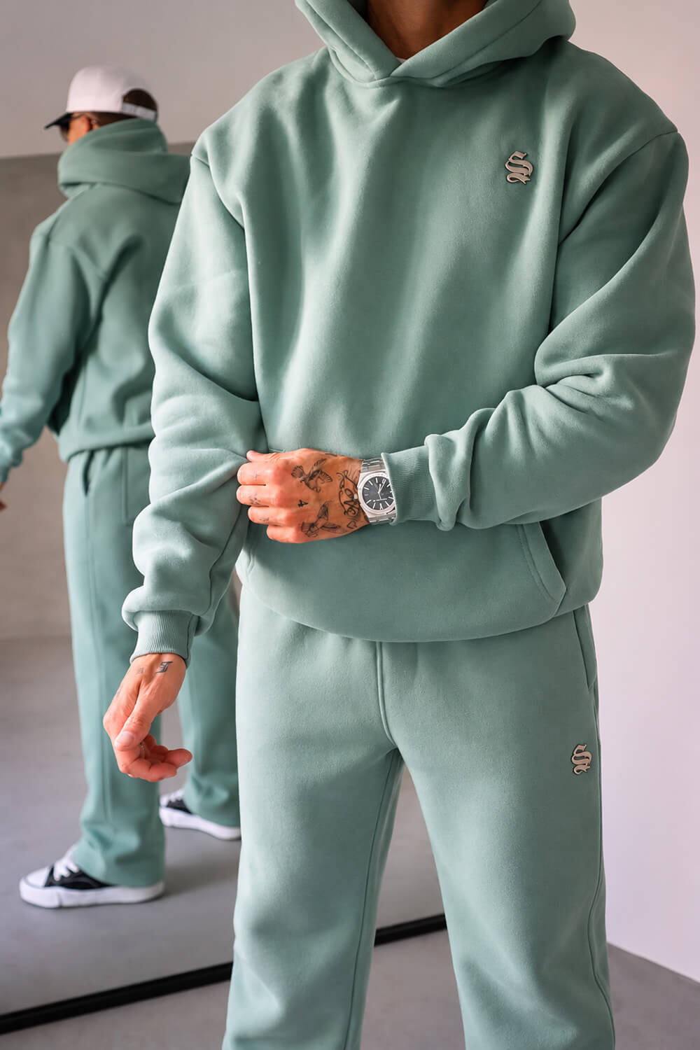 A person wearing a matching sage green hoodie and sweatpants set, with a small embroidered logo on the chest and left thigh. The image includes a mirror reflection in the background showing the full outfit.