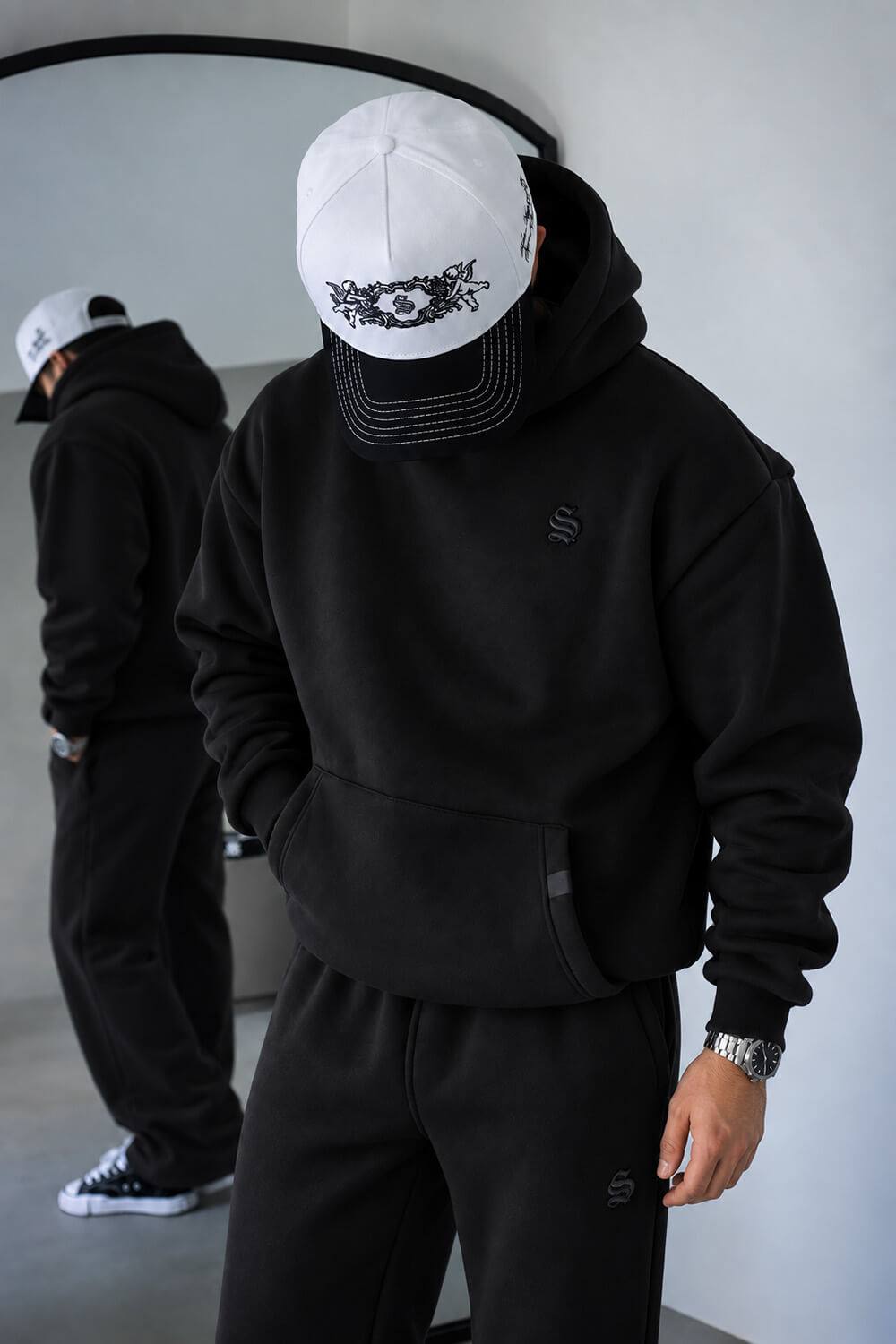 A person wearing a black pullover hoodie, black sweatpants, and a white baseball cap with a black graphic logo, standing in front of a mirror that reflects their back. The setting is a minimalist indoor space with a light gray wall.