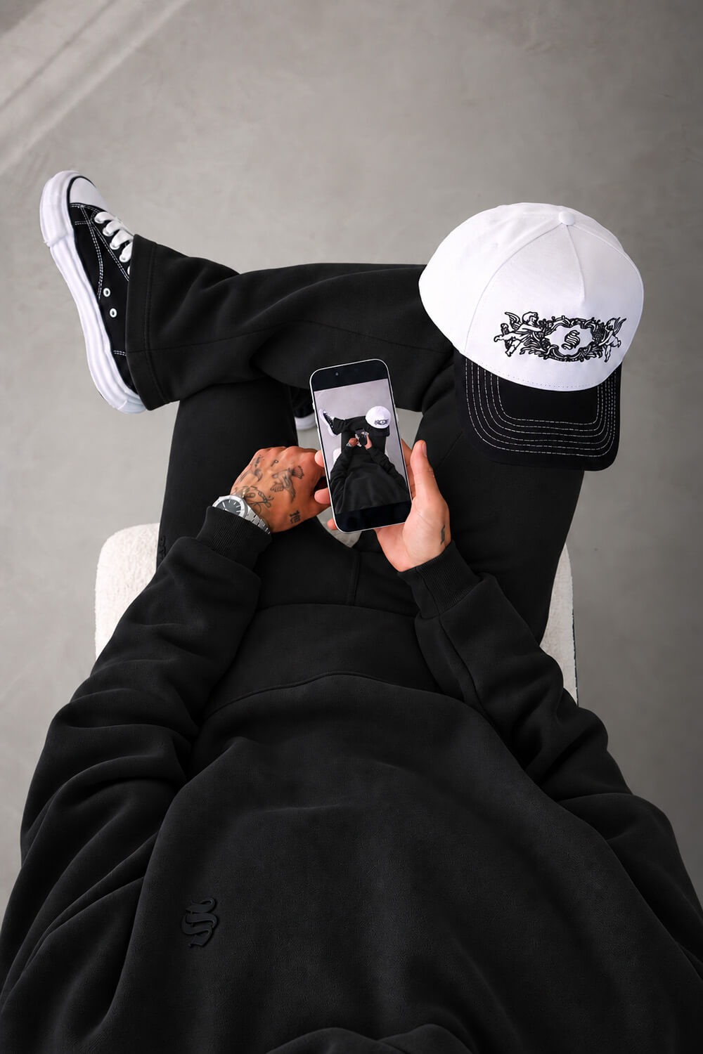 A person wearing a black hoodie, black sweatpants, a white and black baseball cap, and black Converse sneakers, lying down and holding a smartphone.