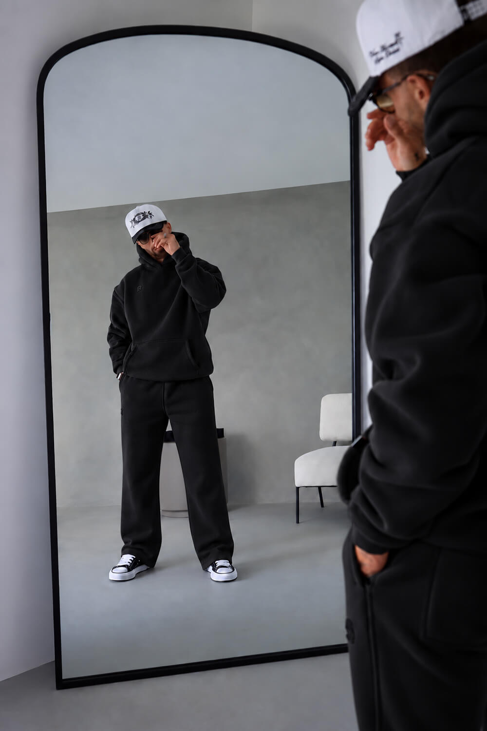 A person in a black hoodie and black sweatpants stands in front of a large floor-to-ceiling mirror, looking at their reflection while wearing a white cap. The room has a minimalist gray concrete wall and floor, with a white chair visible in the background.