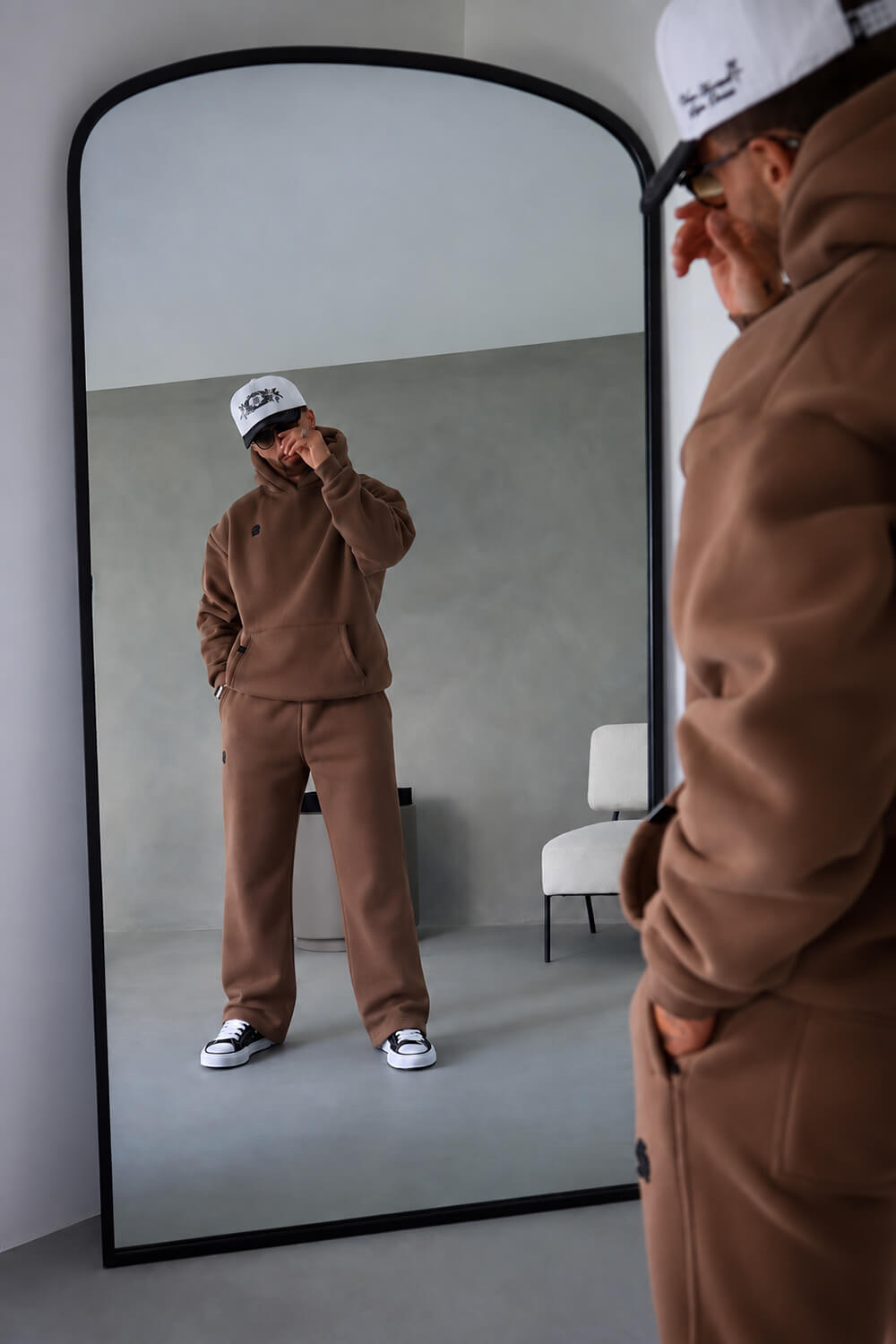 A person wearing a matching brown sweatshirt and sweatpants set stands in front of a large floor mirror, with their reflection visible. The person is also wearing a white cap and black-and-white sneakers.