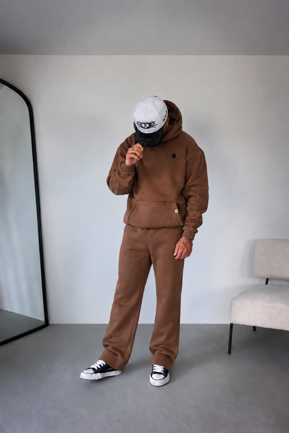 A person modeling a matching brown hoodie and sweatpants set, wearing a white cap and black-and-white sneakers, standing in a minimalist indoor room with a mirror and chair.