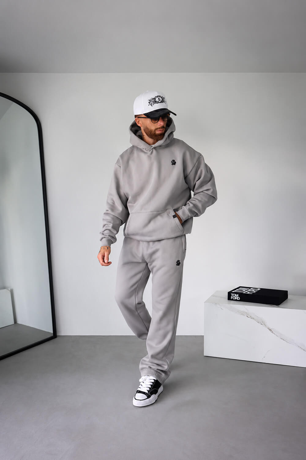 A man modeling a light gray two-piece tracksuit consisting of a pullover hoodie and matching sweatpants, styled with a white cap, sunglasses, and black-and-white sneakers in a minimalist room.