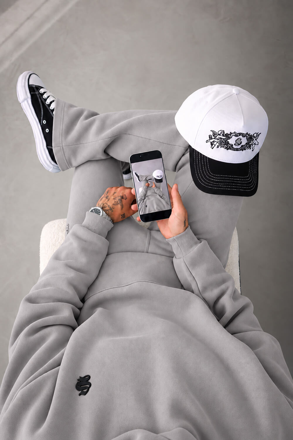 A person wearing a light gray sweatshirt and matching sweatpants, black high-top sneakers, and a white cap with a black graphic, lying down and holding a smartphone.