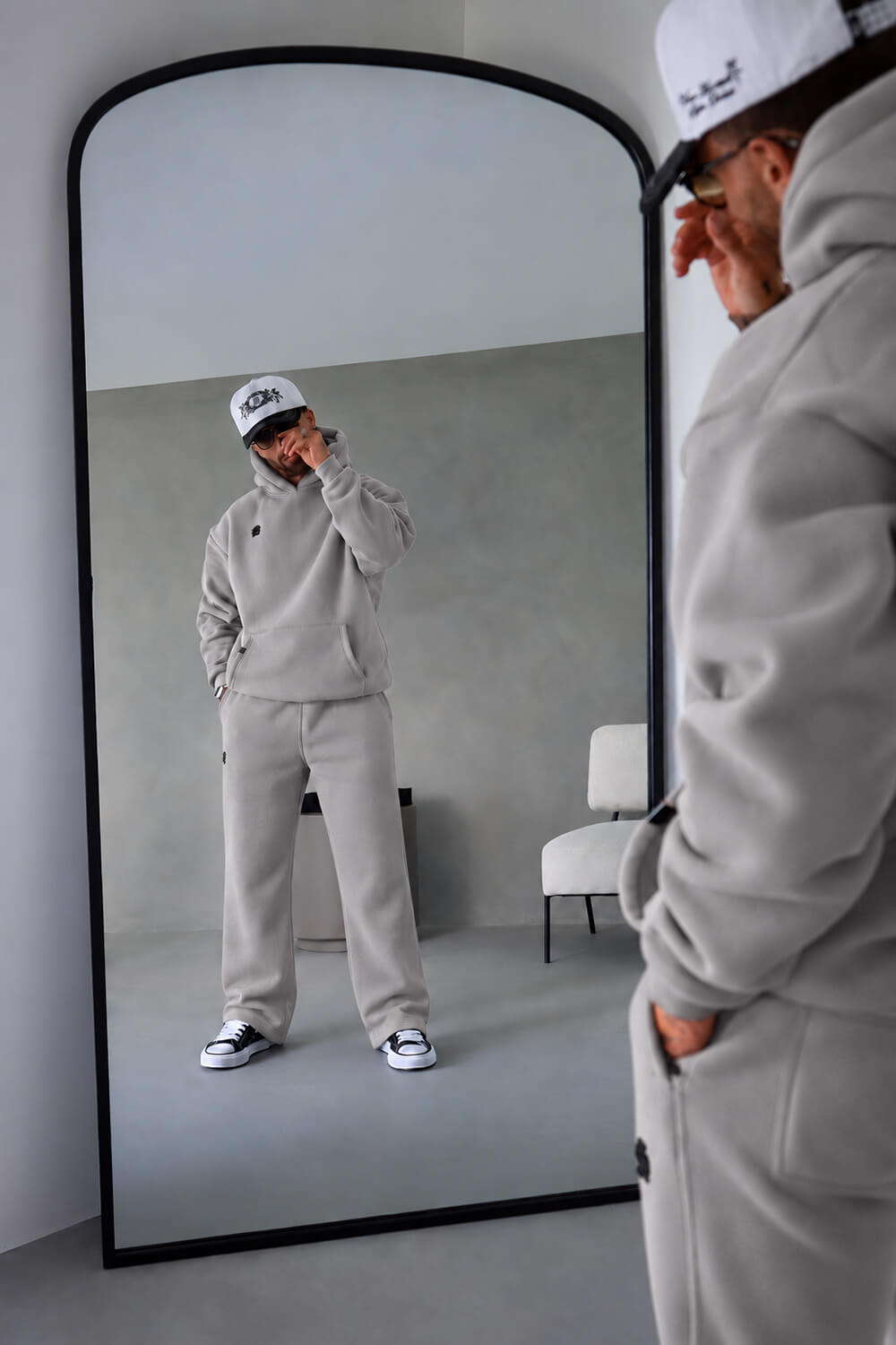 A person wearing a light gray sweatshirt and matching sweatpants stands in front of a large floor mirror, with their reflection visible. The person is also wearing a white cap and black-and-white sneakers.