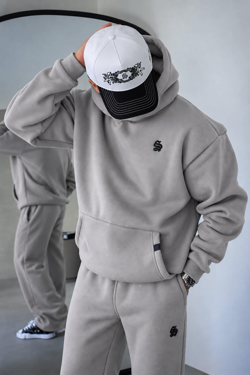 A person wearing a light gray matching hoodie and sweatpants set with small black embroidered logos, paired with a white and black baseball cap, standing indoors with a mirror reflecting their back.