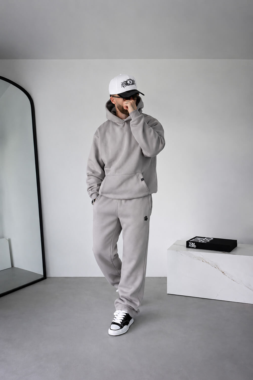 A man wearing a light gray two-piece tracksuit with a matching hoodie and sweatpants, a white baseball cap, and black-and-white sneakers, standing in a minimalist gray room.