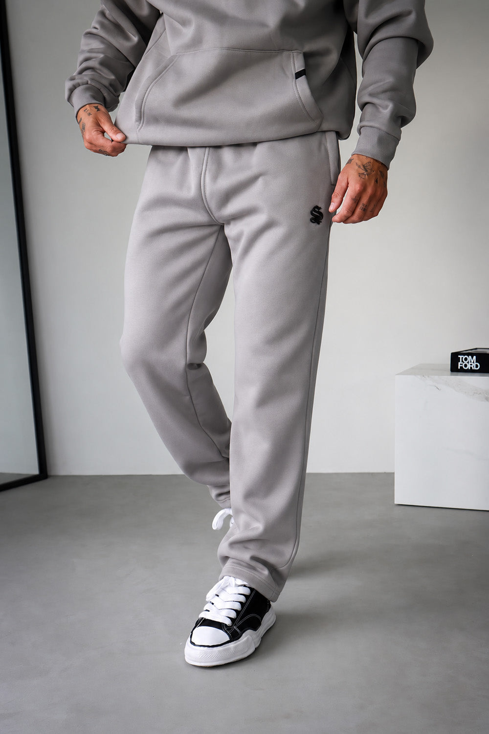 A person wearing light gray jogger sweatpants with a small black embroidered logo on the left thigh, paired with a matching gray hoodie and black-and-white sneakers, standing in a minimalist studio setting.